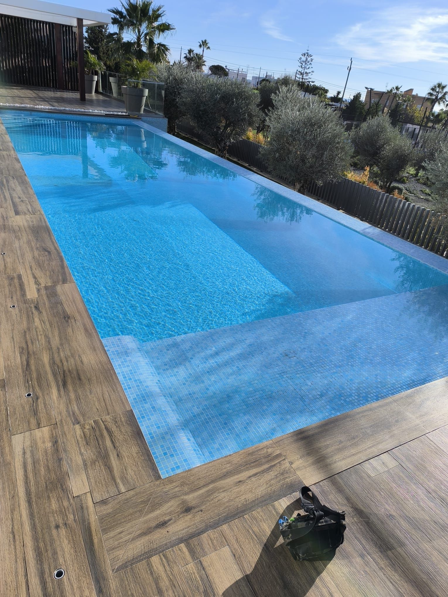 Una piscina infinita rectangular con agua azul y terraza de madera, con vistas a un jardín.