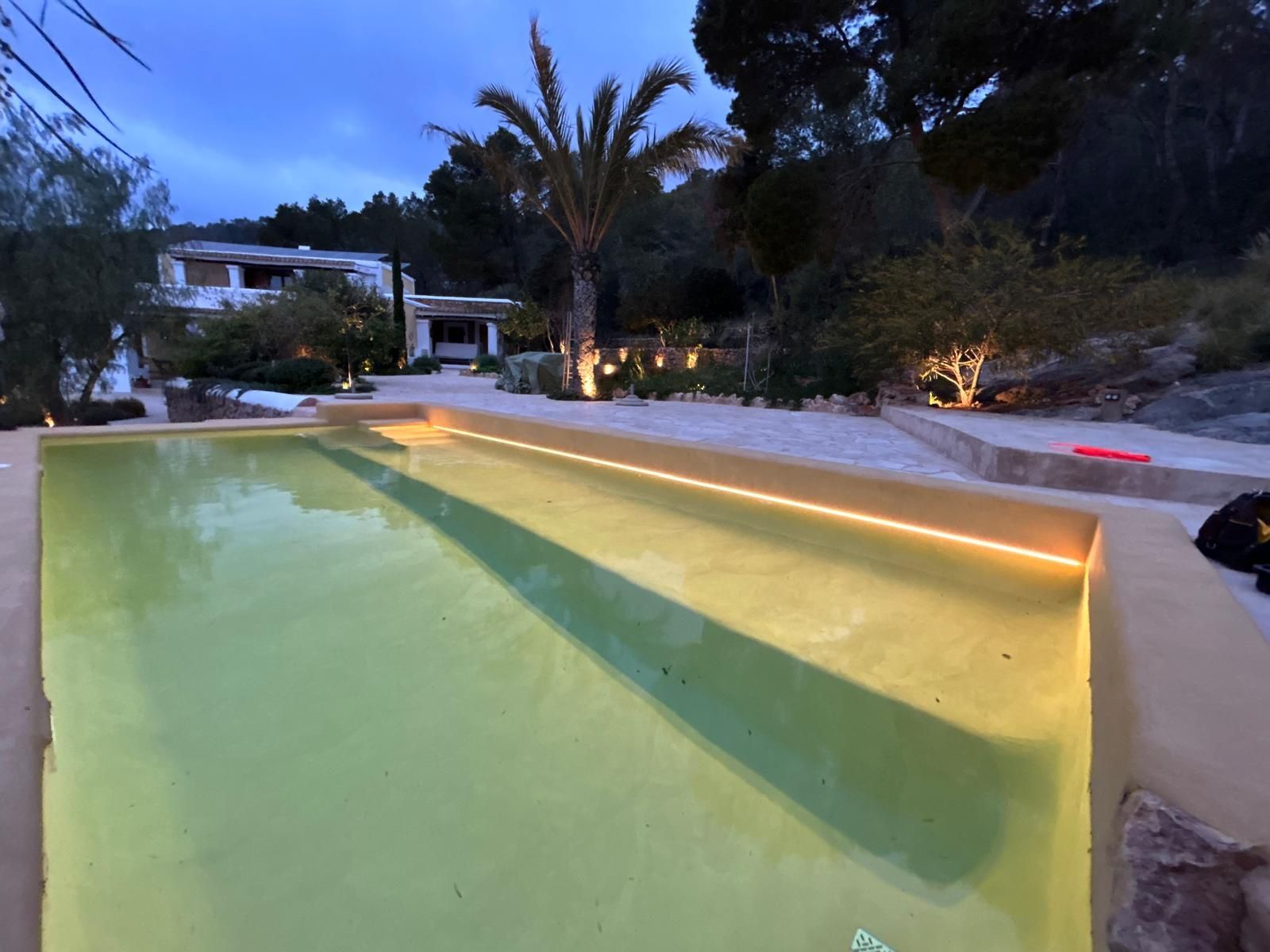 Piscina iluminada con luces amarillas al anochecer, rodeada de vegetación y un edificio blanco.