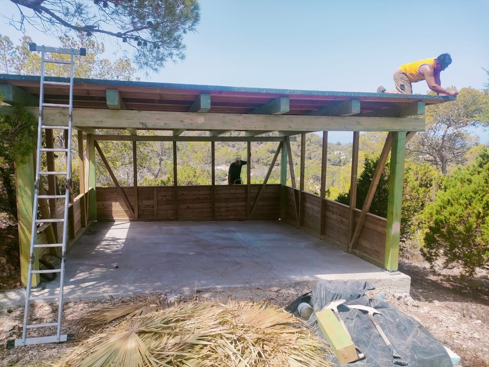 Hombres construyendo una estructura de madera al aire libre. Madera pintada de verde, cielo azul y sol radiante.