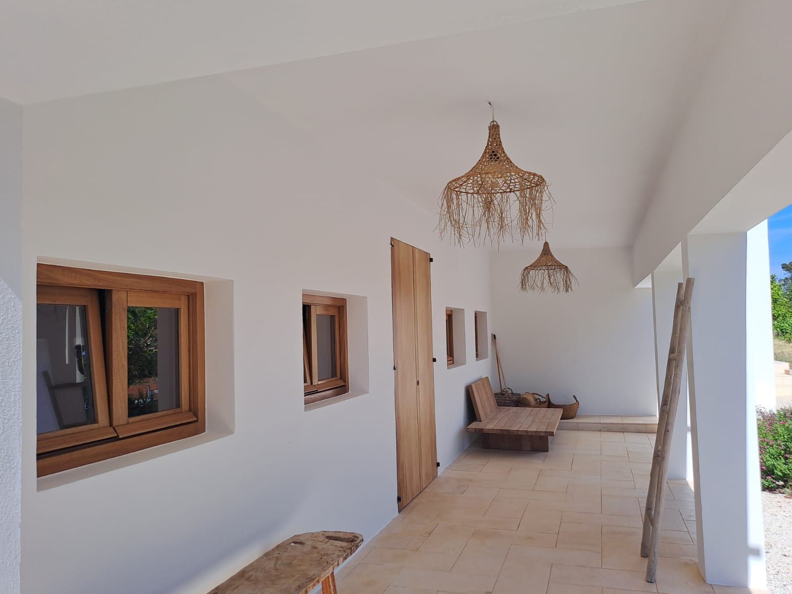 Veranda blanca con puerta de madera, ventanas y lámparas colgantes tejidas.