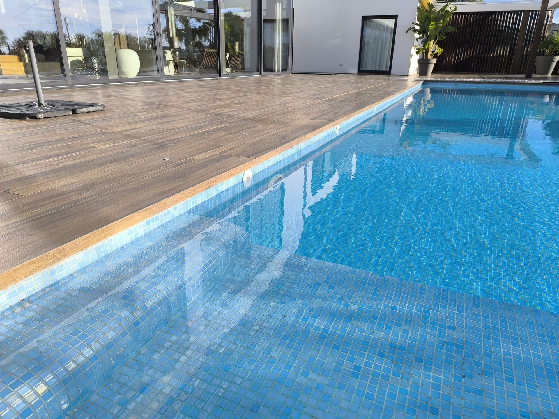 Piscina de agua azul e interior de azulejos, junto a tarima de madera y una casa moderna.