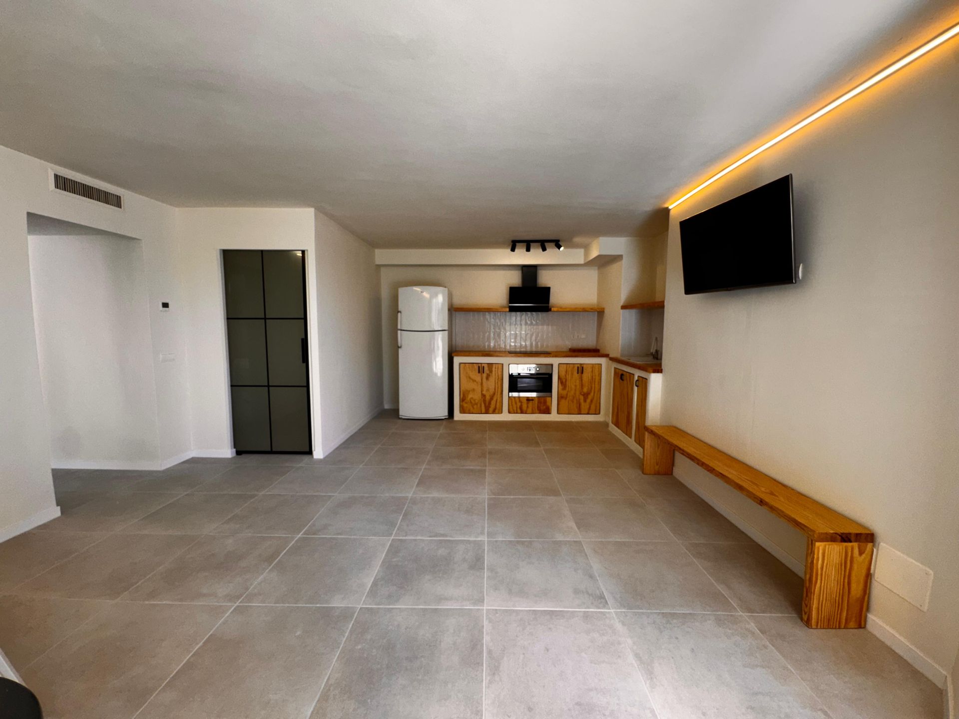 Apartamento de concepto abierto con suelo de baldosas, cocina y TV empotrada. Muebles y banco de madera clara.