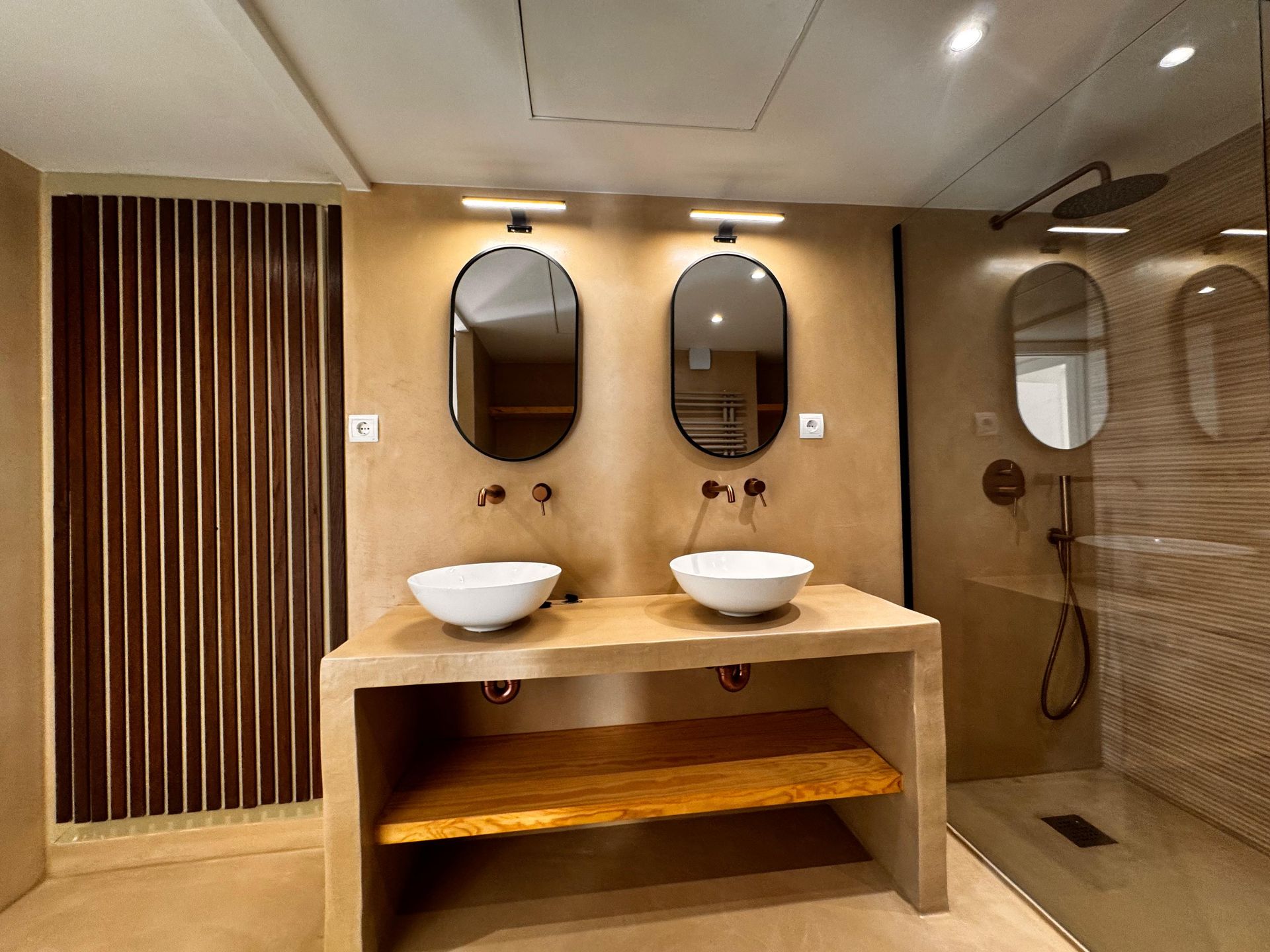 Baño con espejos ovalados, lavabos sobre encimera y estanterías de madera; ducha a la derecha, lamas verticales de madera a la izquierda.