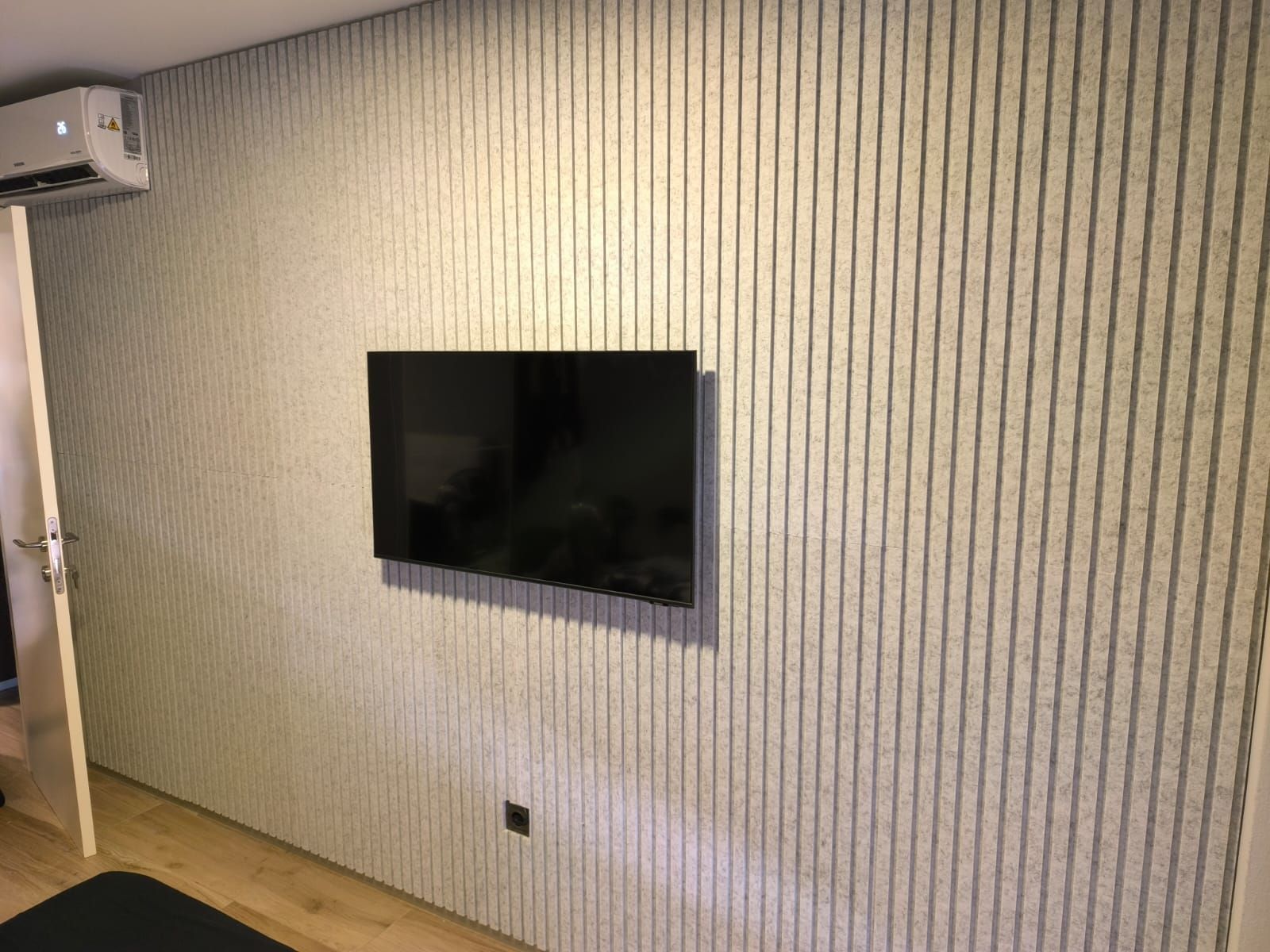 Un televisor montado en una pared con nervaduras verticales, con un aparato de aire acondicionado encima y una puerta a la izquierda.