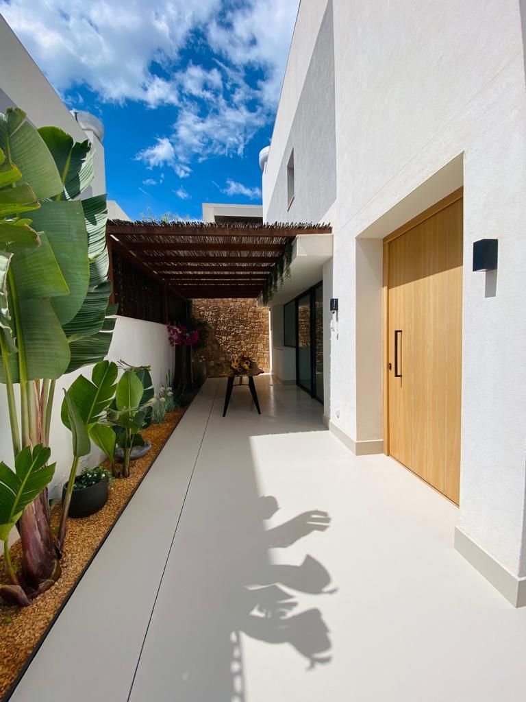 Casa moderna blanca con puerta de madera y pasarela sombreada, cielo azul brillante.
