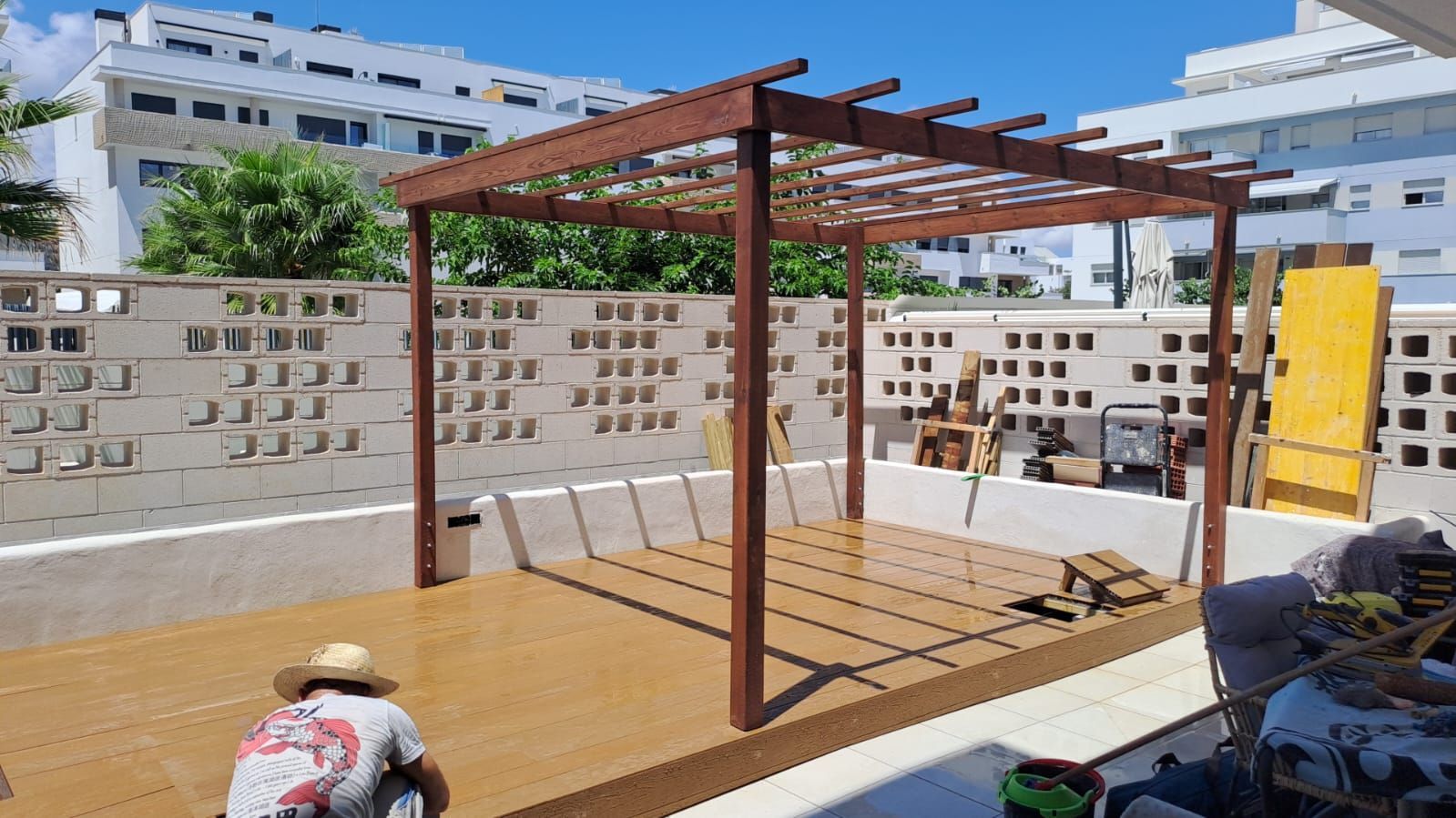 Una persona trabaja en una terraza de madera con pérgola, junto a una pared de recortes cuadrados, en un patio.