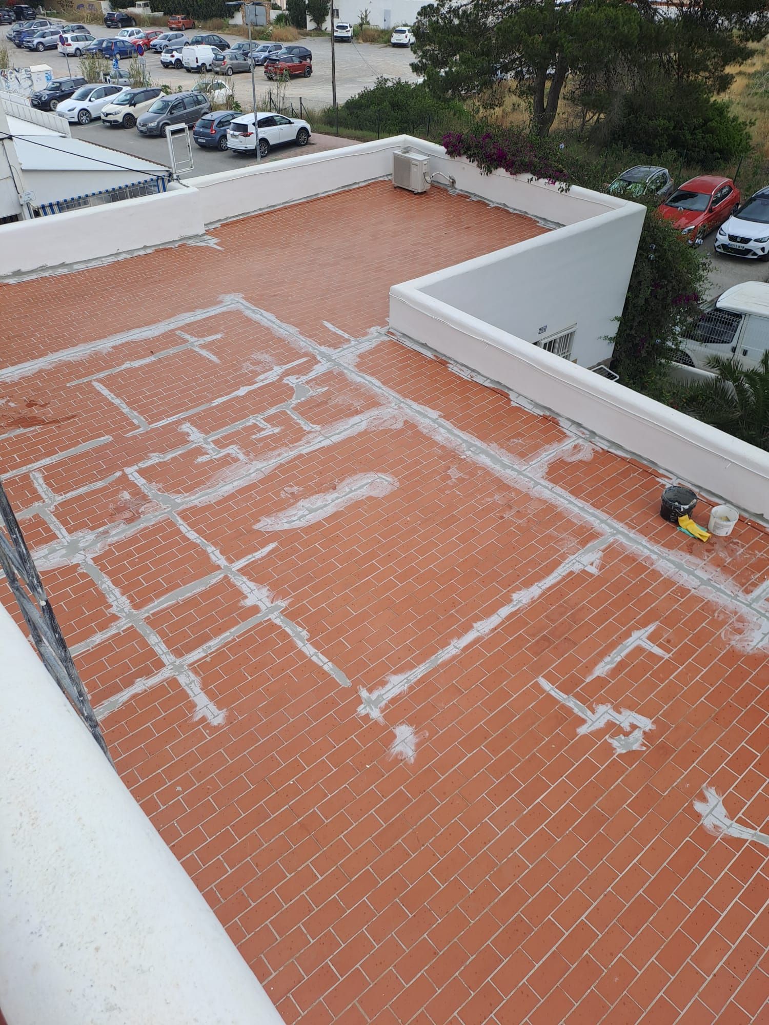 Techo de tejas rojas con sellador gris, visto desde arriba. Coches y edificios visibles al fondo.