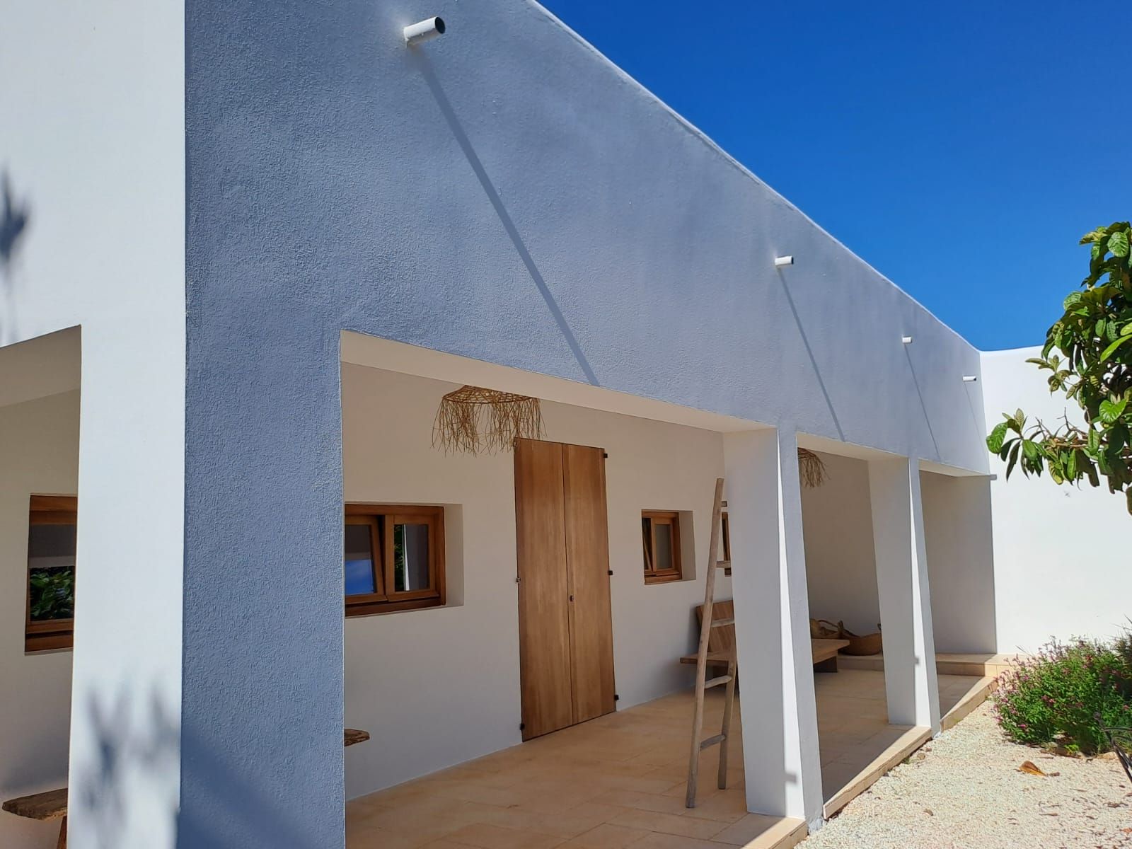 Edificio blanco con puerta y ventanas de madera, patio cubierto, cielo azul.
