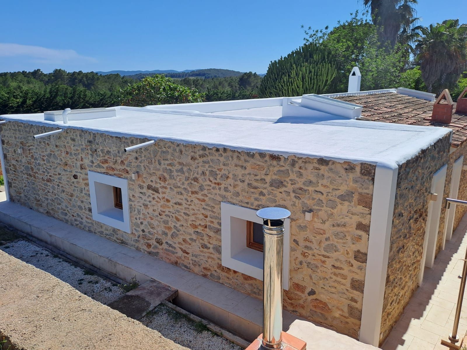 Casa de piedra con ventanas revestidas de blanco y tejado plano blanco, en un entorno exterior soleado.