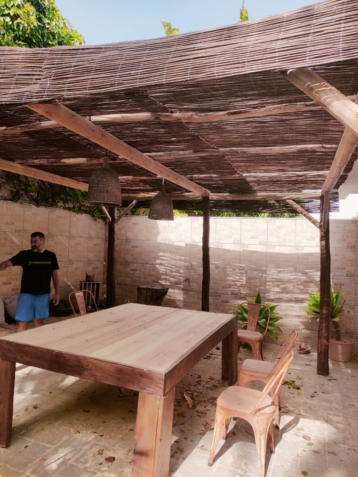 Pérgola de madera sobre un patio con mesa, sillas y un hombre haciendo gestos.
