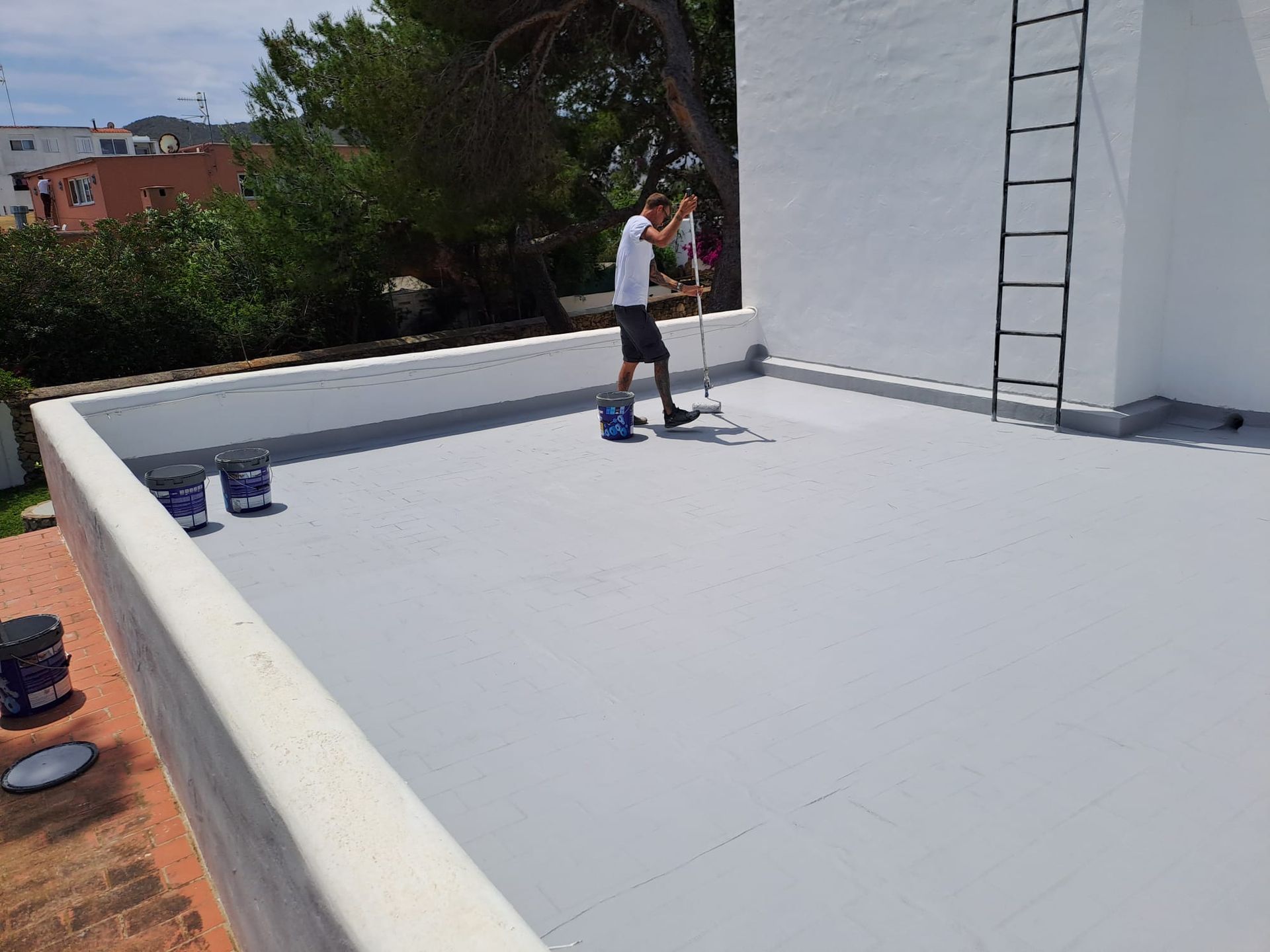 Hombre pintando de blanco un tejado plano y gris. Se ven cubos de pintura y una escalera.