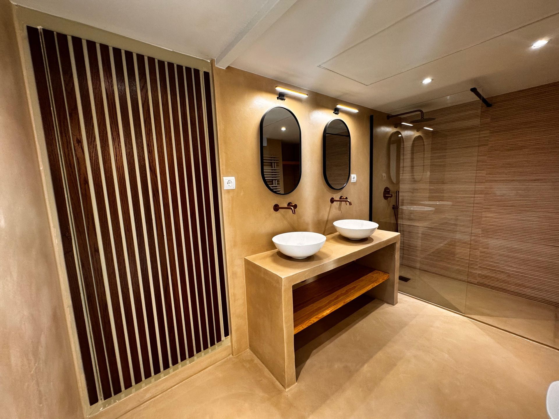Baño con dos lavabos, dos espejos ovalados y ducha a ras de suelo. Panel de pared con listones de madera. Paredes beige.