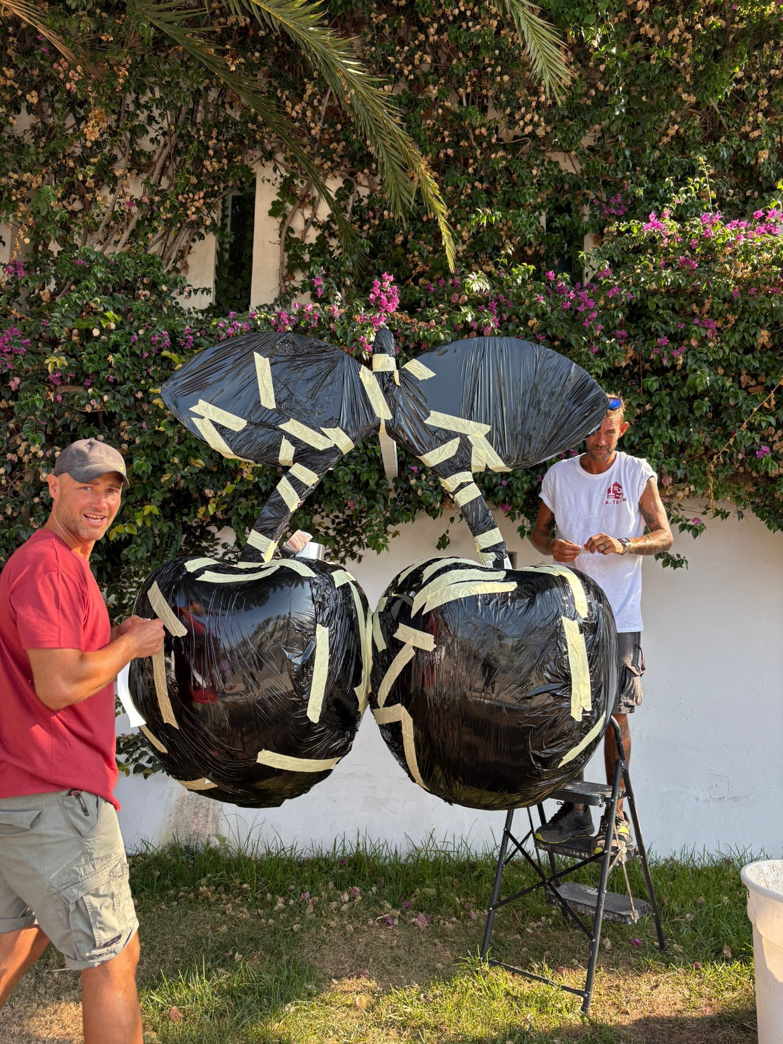 Dos hombres arreglan una escultura gigante de cerezo negro con cinta color canela al aire libre frente a unos arbustos.