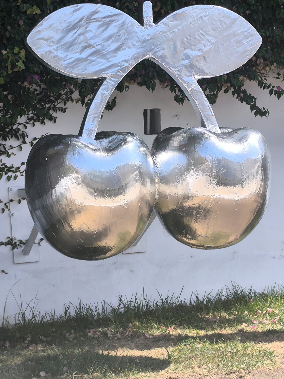 Escultura gigante de cereza plateada contra una pared blanca con vegetación en el fondo.