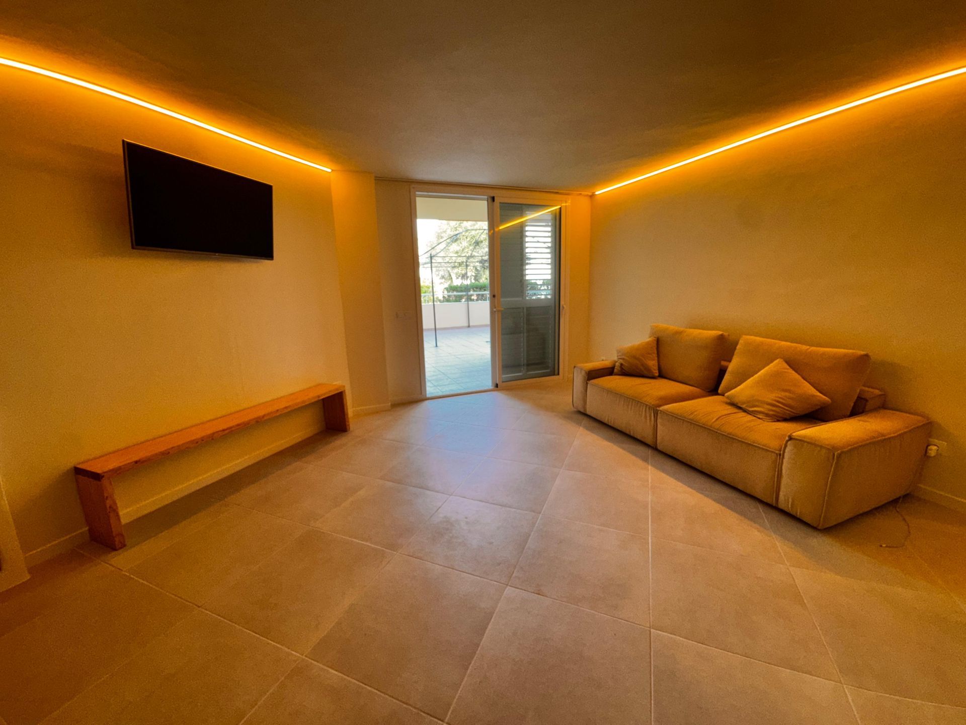 Acogedora sala de estar con iluminación amarilla, sofá, banco, TV y puerta abierta al balcón.