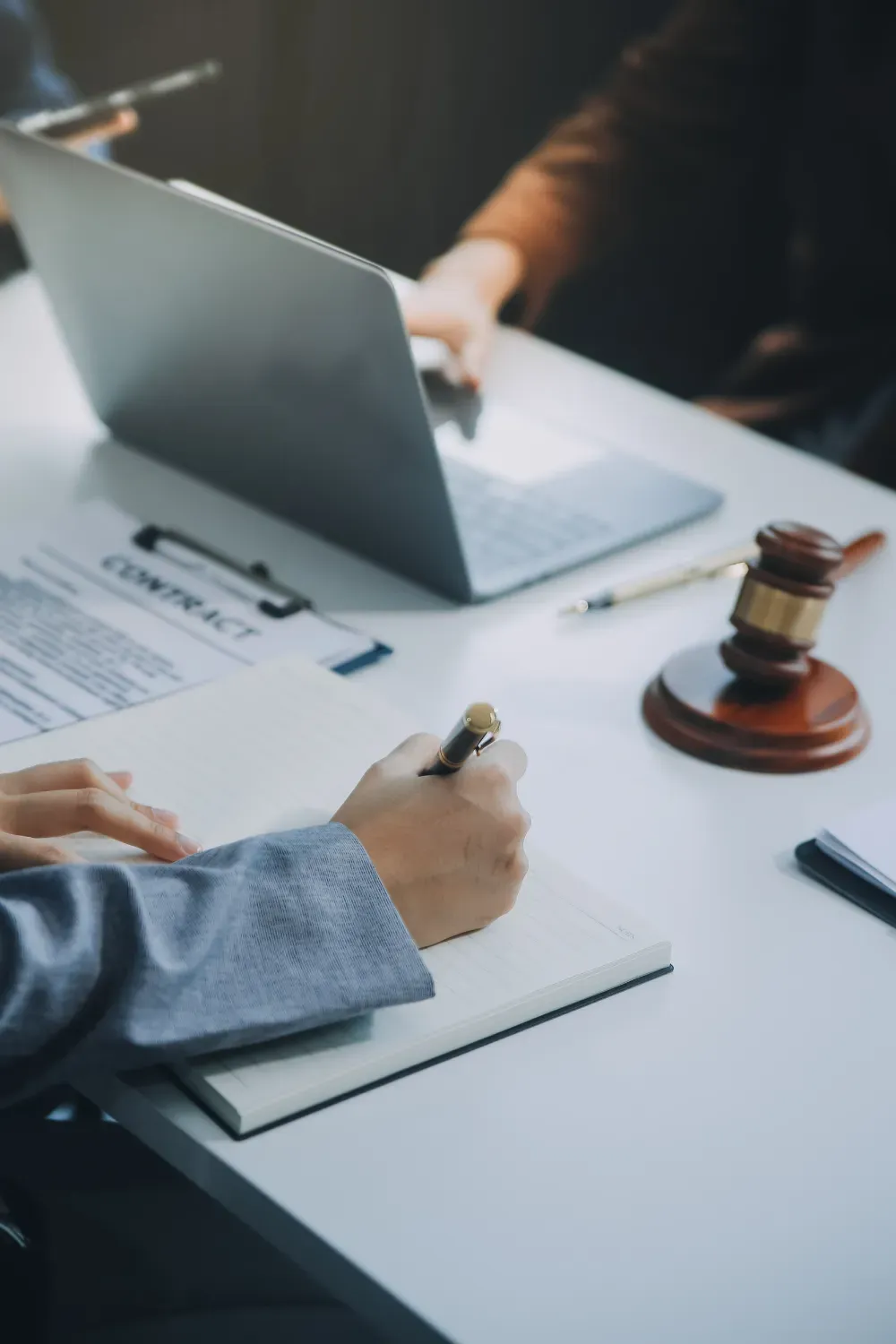 Manos escribiendo notas, computadora portátil, contrato, mazo, bolígrafo sobre una mesa blanca.