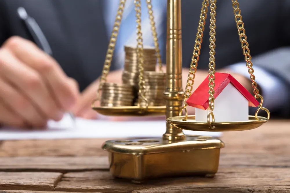 Una balanza con monedas en un lado y una casa en el otro; una persona escribe en el fondo.