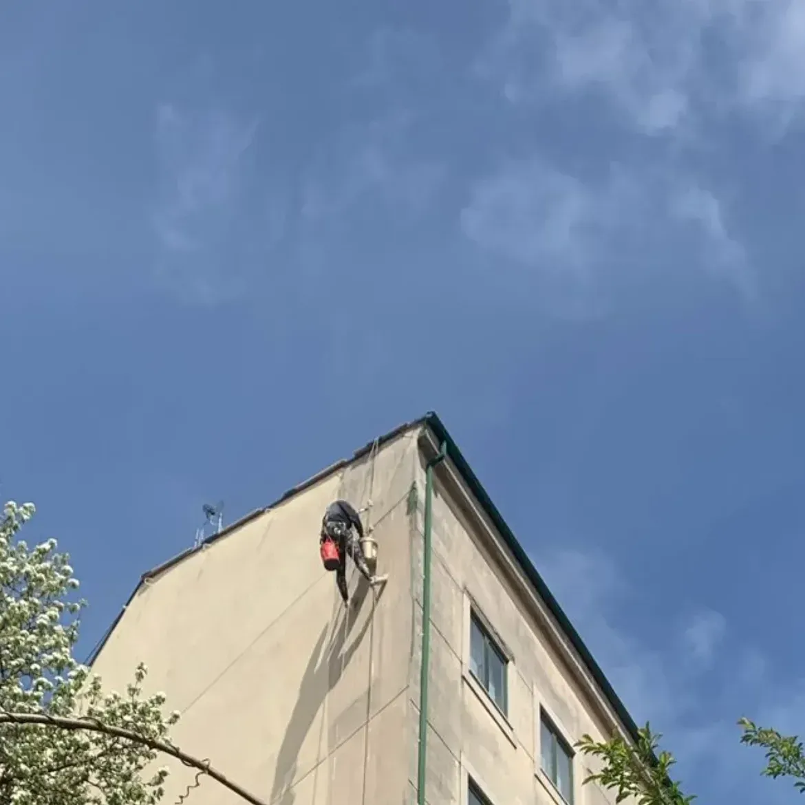 La esquina de un edificio con un limpiador de ventanas suspendido de cuerdas contra un cielo azul.
