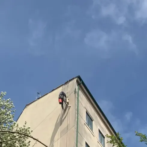 La esquina de un edificio con un limpiador de ventanas suspendido de cuerdas contra un cielo azul.