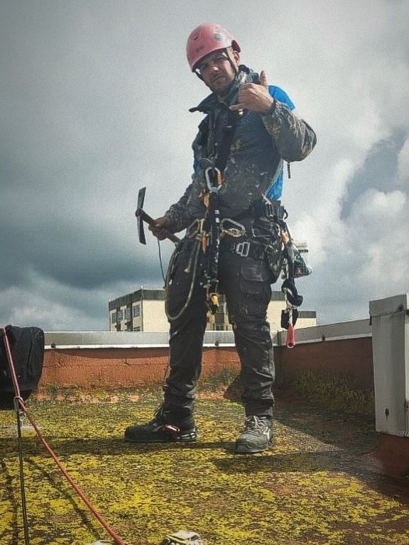 Un techador con casco rosa y arnés azul está de pie en un tejado mojado, sosteniendo herramientas bajo un cielo nublado.