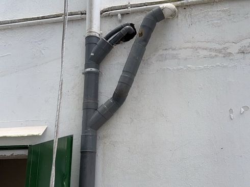 Tuberías de desagüe y conductos de ventilación grises que recorren una pared exterior blanca junto a una puerta verde.