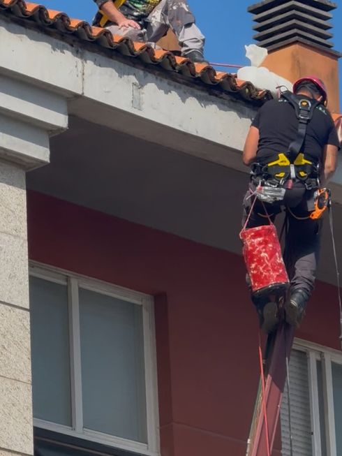 Un trabajador, subido a una escalera, repara una canaleta de techo junto a una casa con paredes blancas y tejas de terracota.