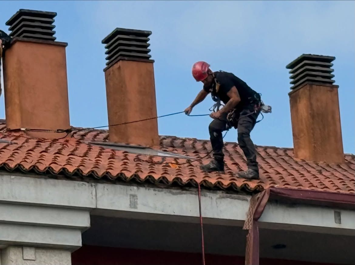 Un trabajador se encuentra en un tejado de tejas rojas, utilizando una cuerda cerca de tres chimeneas bajo un cielo azul.