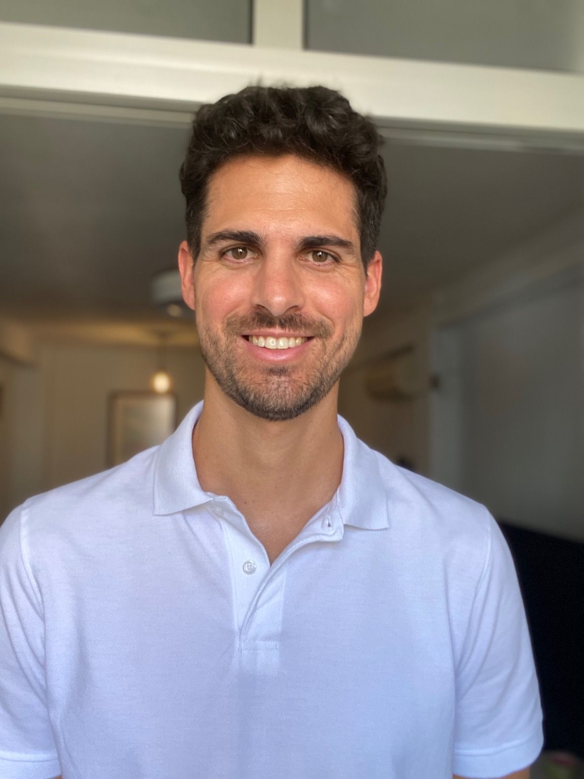 Hombre de cabello oscuro sonriendo, vistiendo una camisa polo blanca, de pie en el interior.