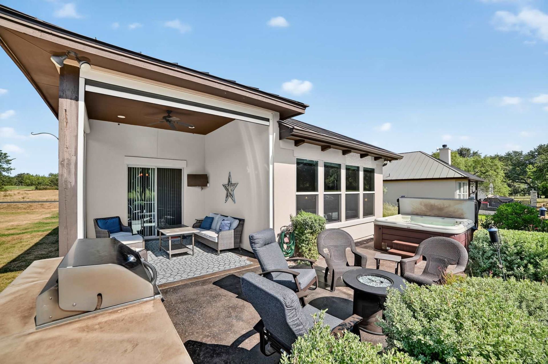 Backyard patio with grill, seating, and hot tub. Beige house with blue sky.