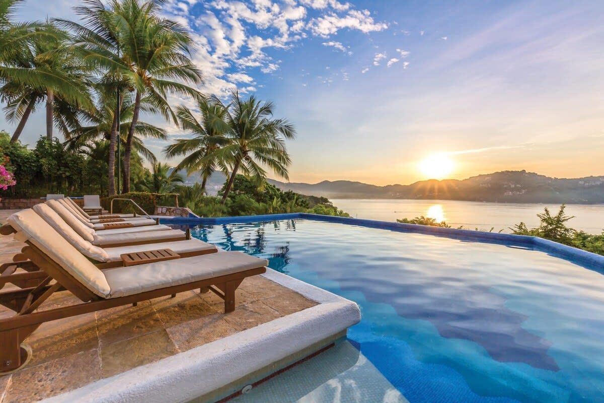 A swimming pool surrounded by chairs and palm trees with a view of the ocean at sunset.