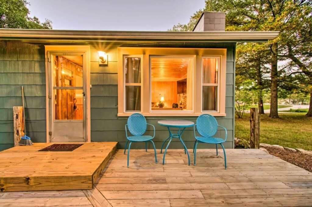 A small teal cabin with a wooden deck; two blue chairs and a table are on the deck.