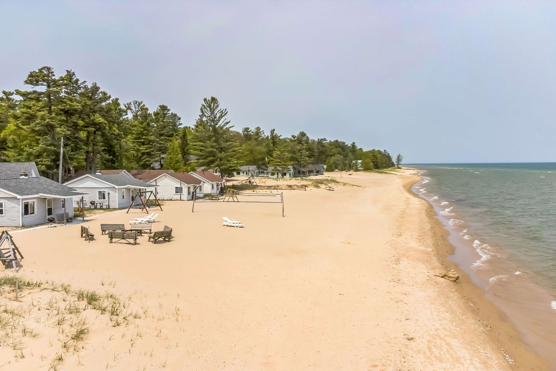 Beachfront cottages on a sandy shore with beach chairs, volleyball net, and calm water under a sunny sky.