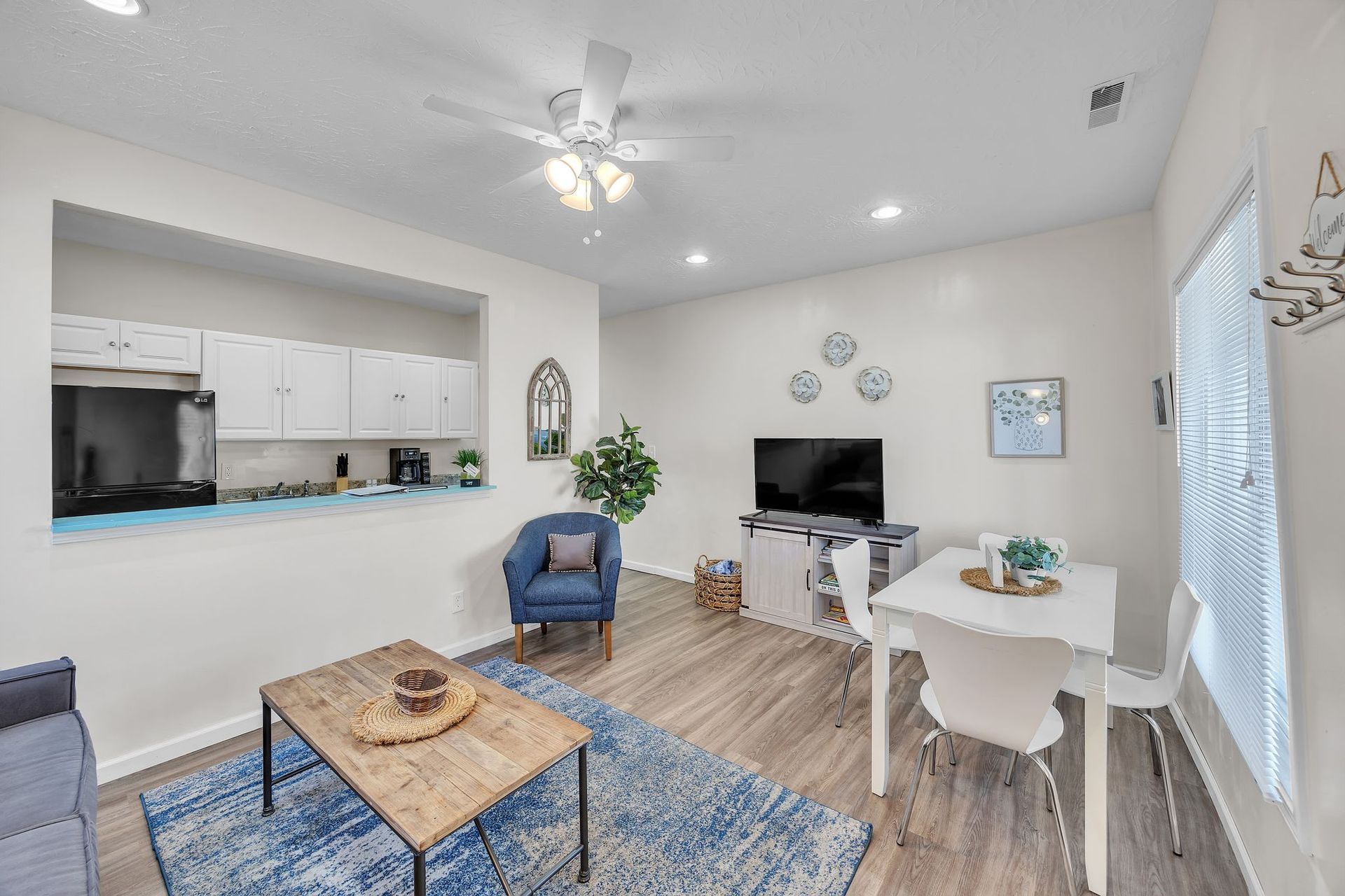 Living room with an open kitchen. Blue rug, wood coffee table, TV, small dining table, and blue chair.