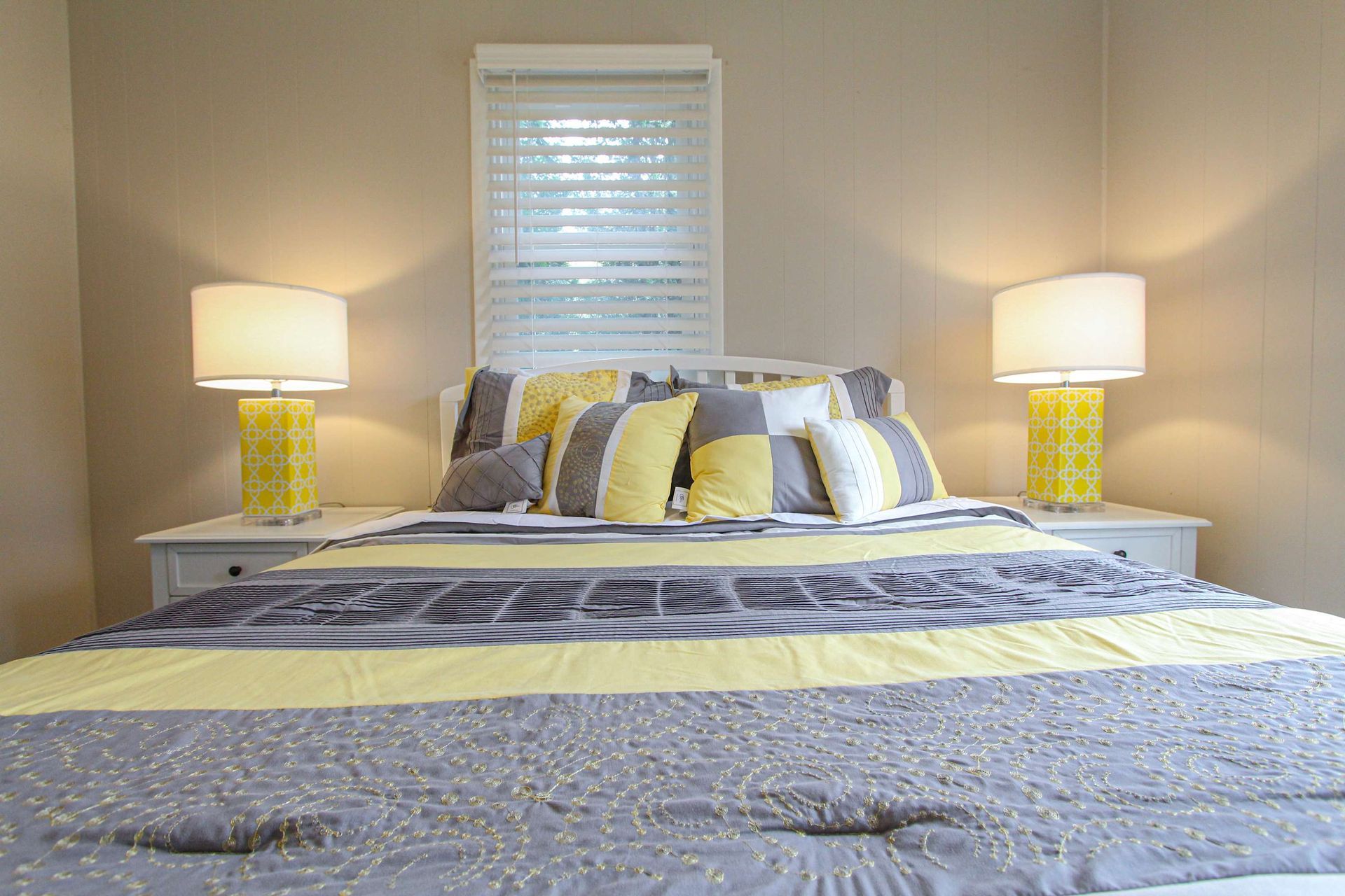 Bedroom with a bed, lamps, pillows, window. Bedding is gray and yellow. Two yellow lamps sit on white nightstands.