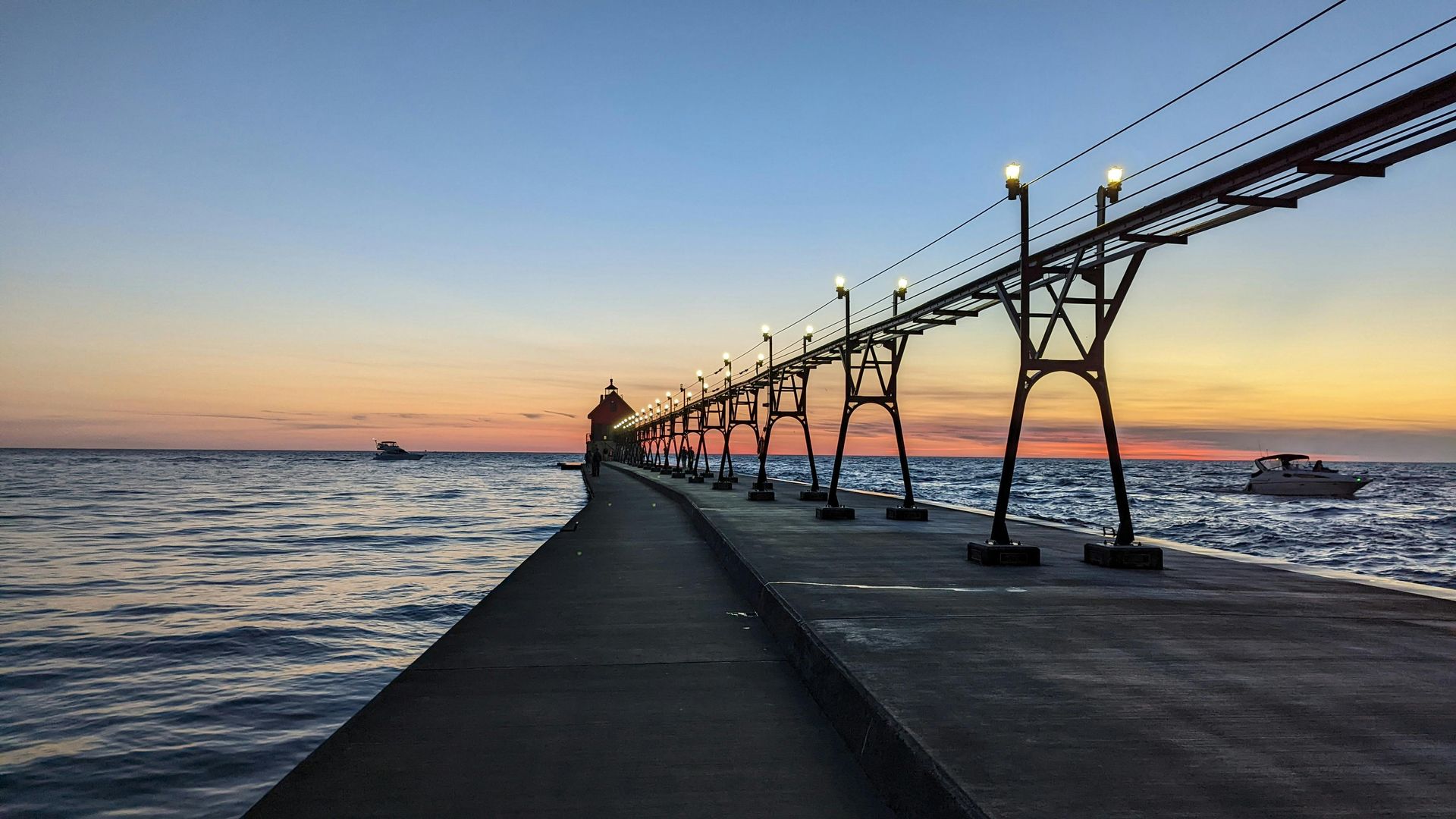 A long pier extends into a body of water at sunset, with lights lining the walkway and two boats nearby.