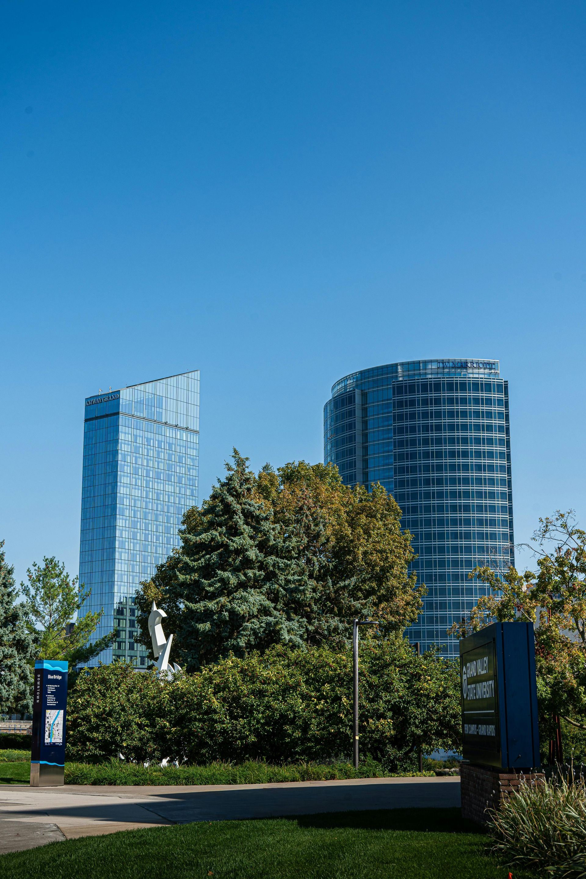 Two modern glass skyscrapers behind a green hedge and park area, under a blue sky.