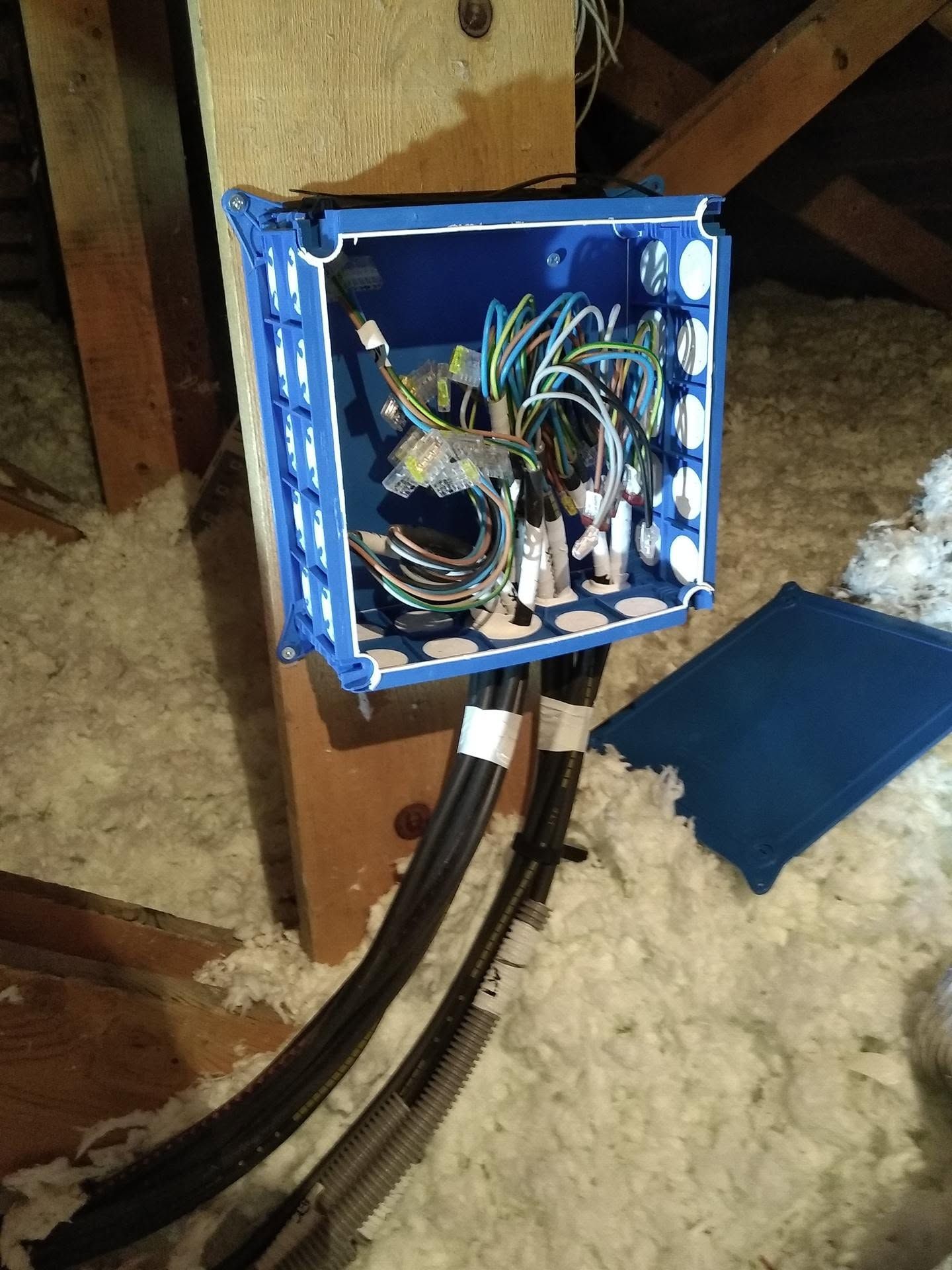 Boîte de dérivation électrique bleue ouverte, avec des fils dénudés, fixée à une poutre en bois dans un grenier.