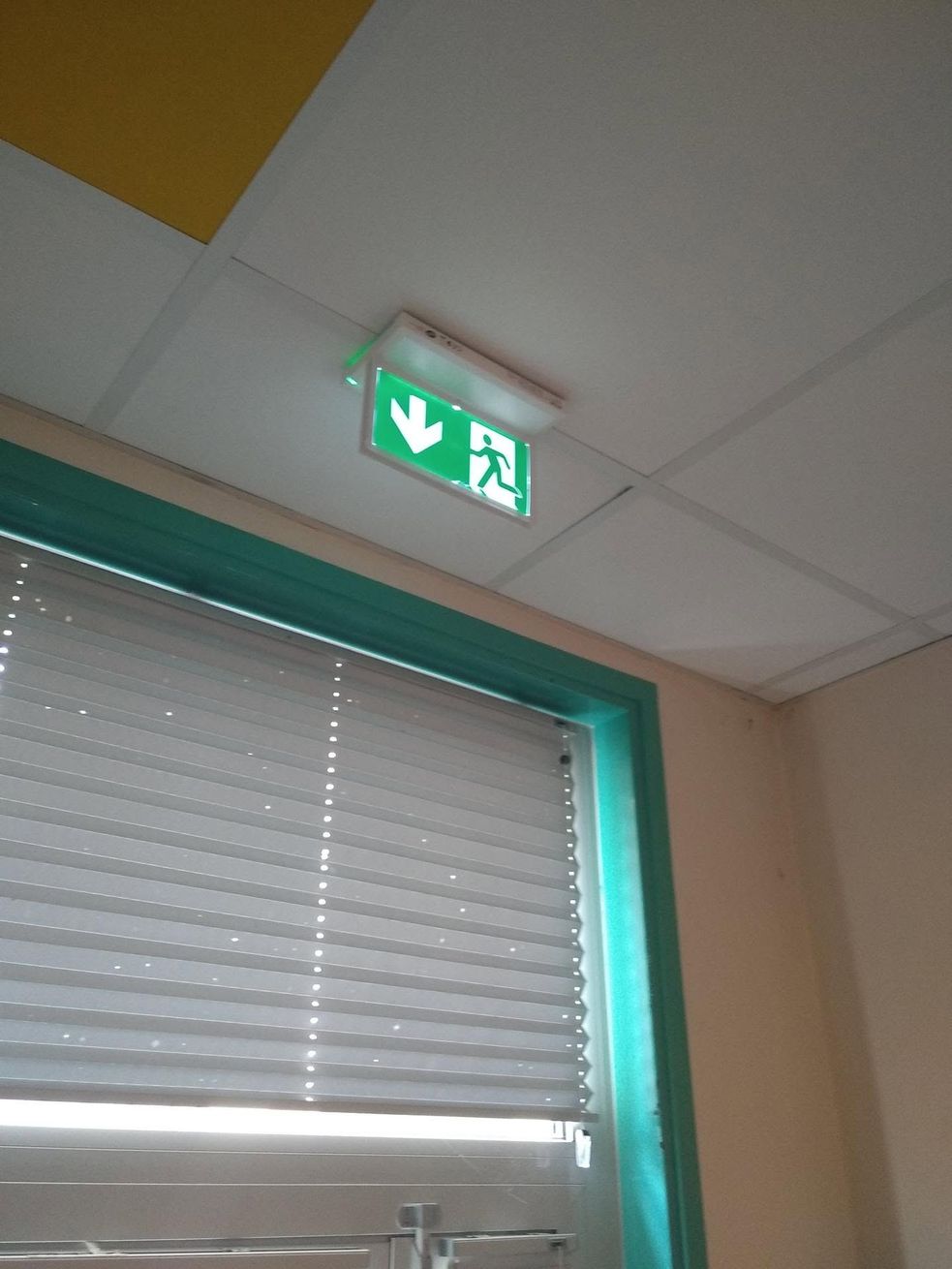 Panneau de sortie de secours vert au-dessus d’une fenêtre aux stores fermés, dans un couloir intérieur