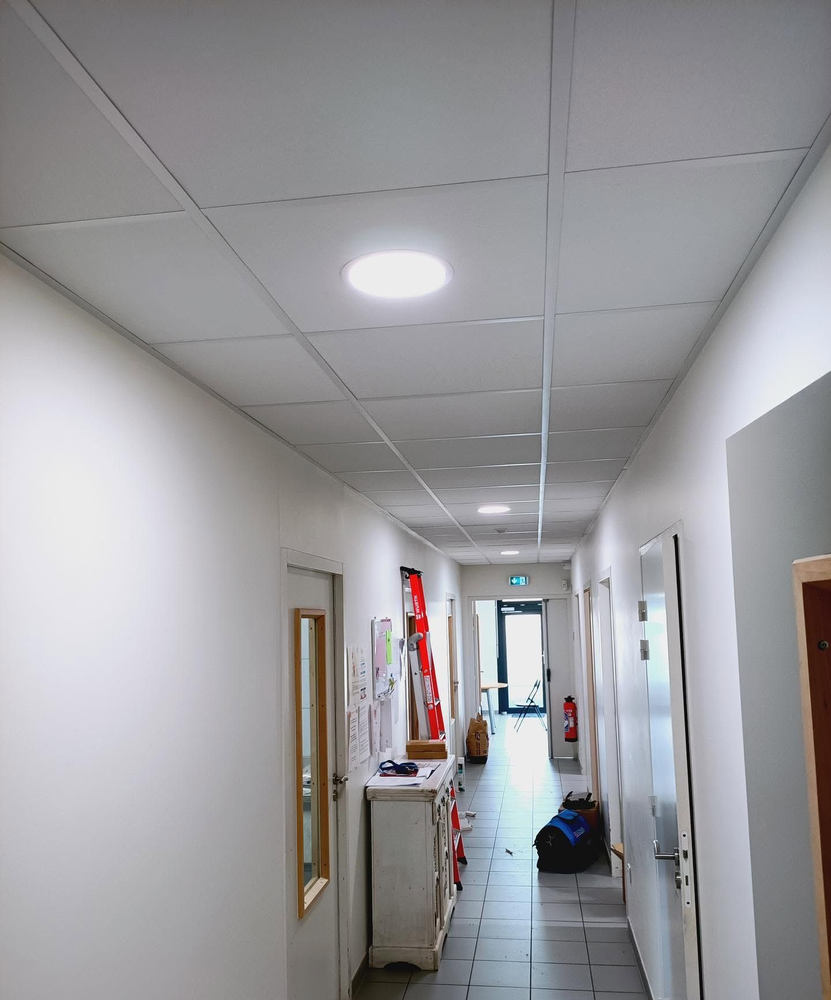 Couloir étroit de bureaux avec un sol carrelé, des murs blancs, des plafonniers et un extincteur rouge