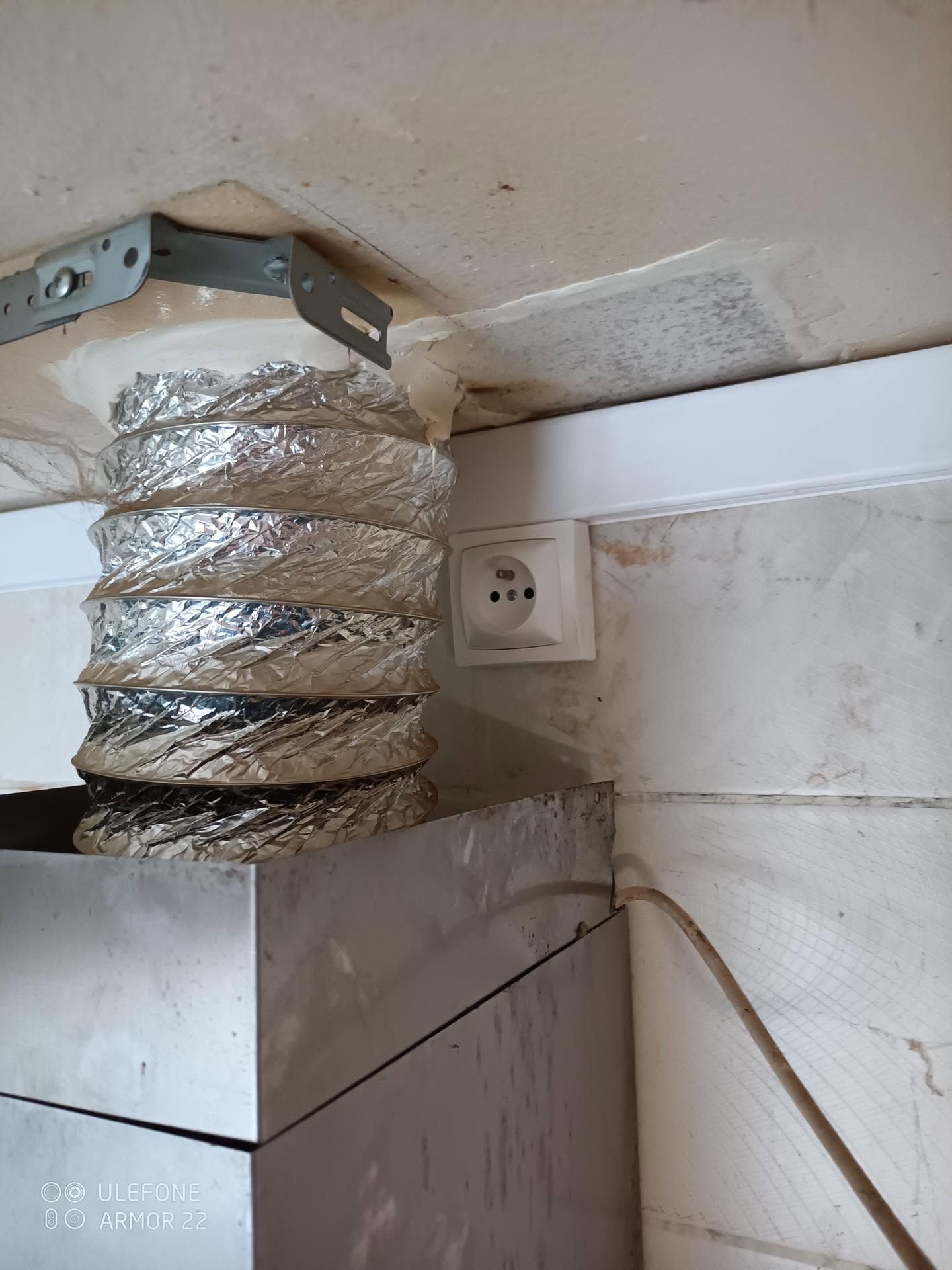 Conduit d’aération flexible argenté raccordé à une ouverture au plafond, à côté d’une prise murale et du dessus d’un meuble.