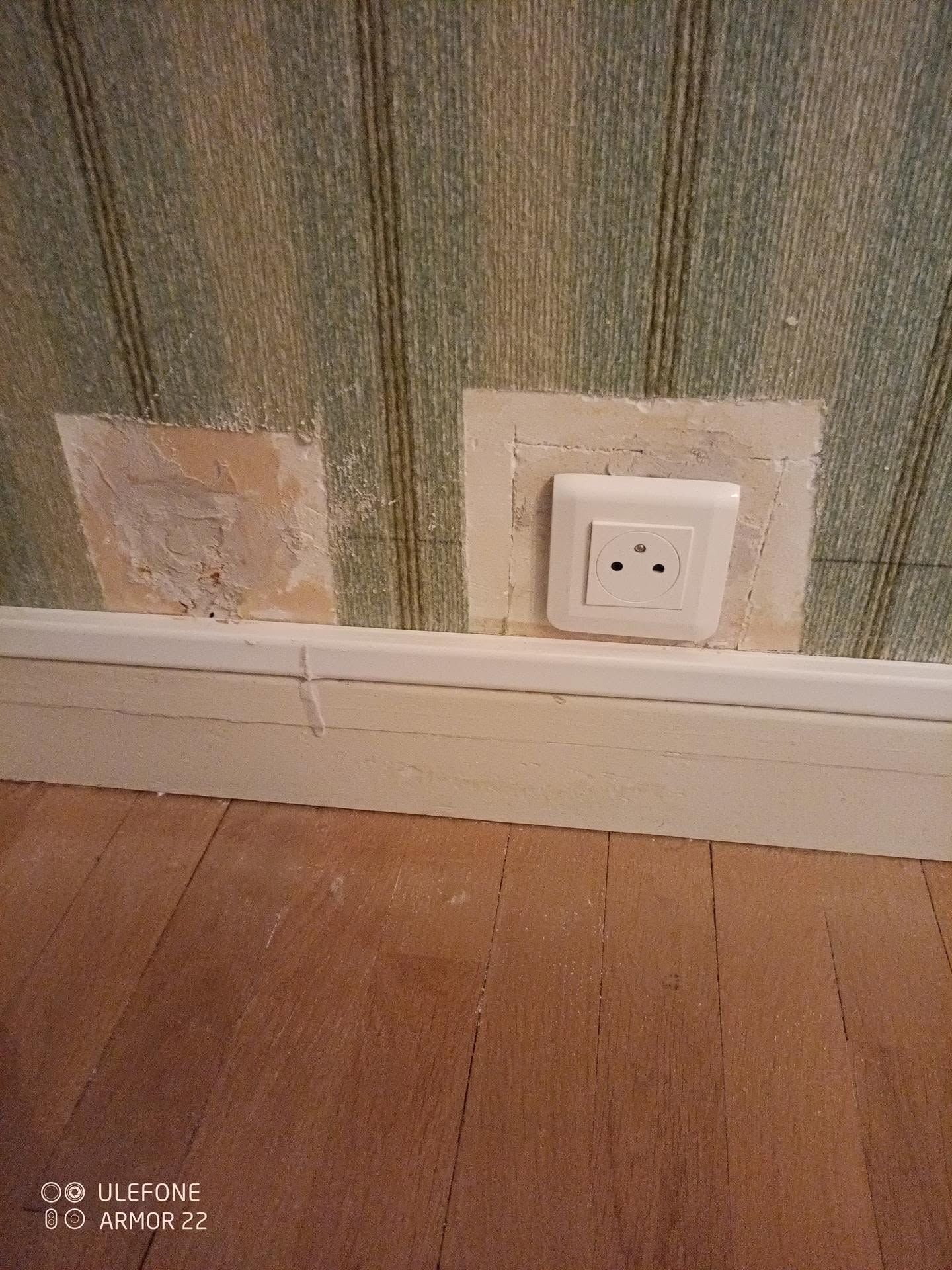Mur avec une prise électrique blanche et des réparations inégales au-dessus d’un plancher en bois et d’une plinthe