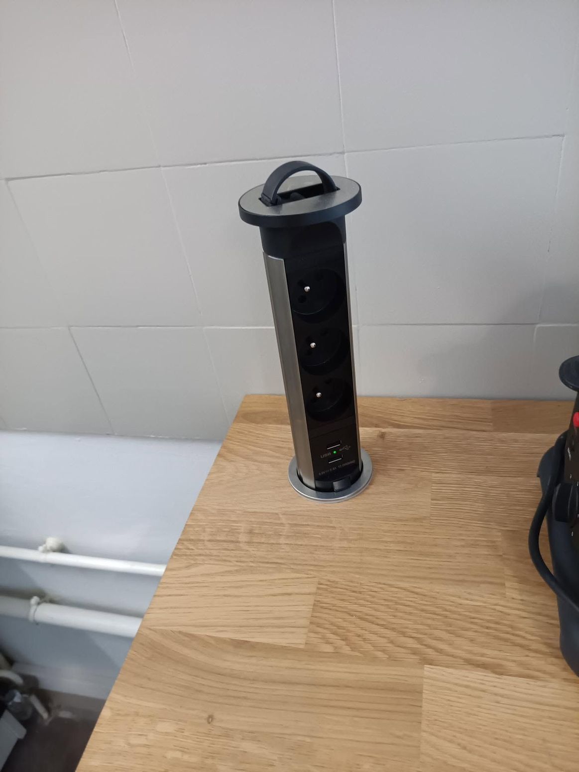 Objet cylindrique noir posé sur un plan de travail en bois, près d’un mur gris et de tuyaux.