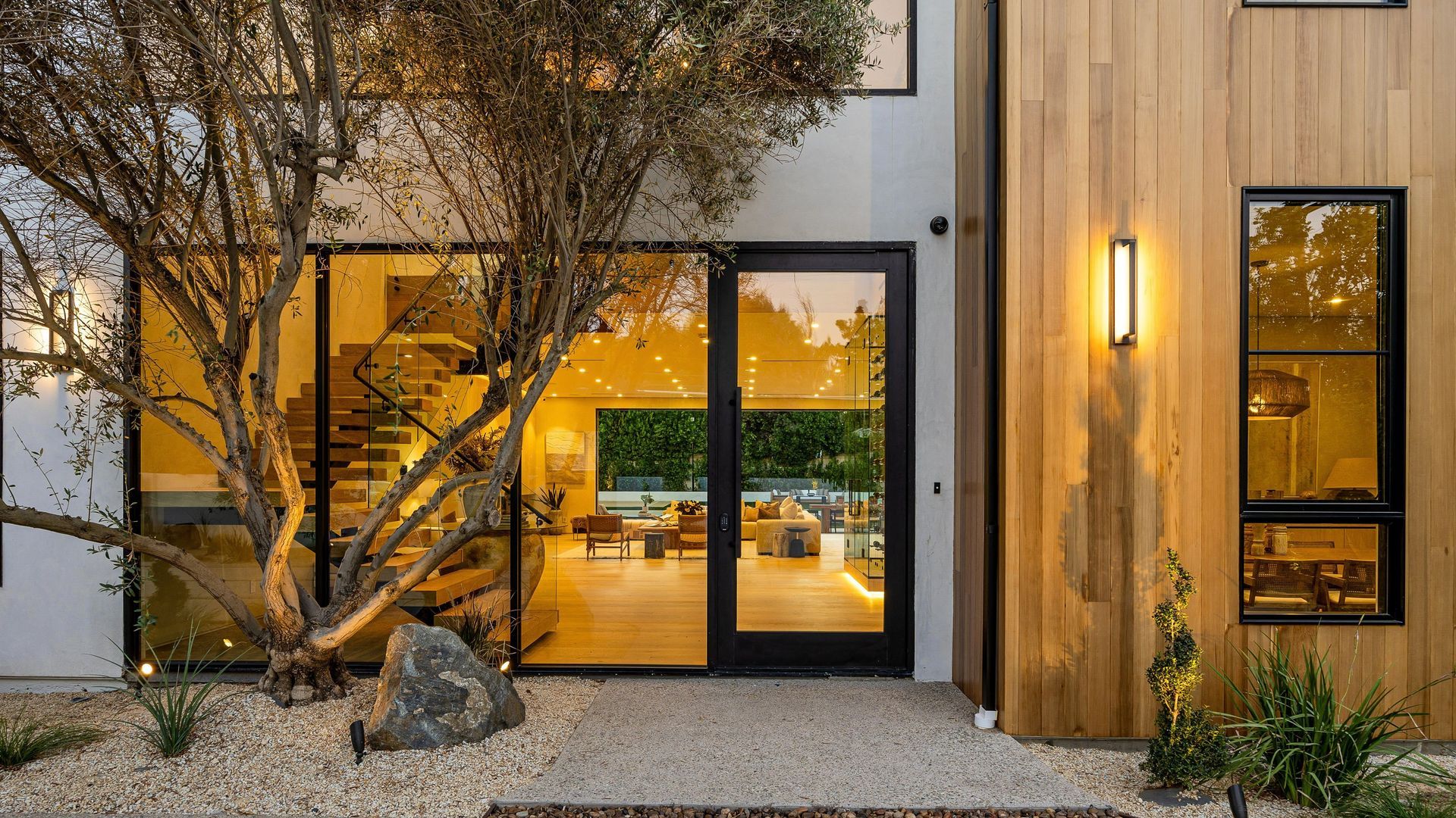 Entrée d’une maison moderne avec des portes vitrées à l’éclairage chaleureux, un bardage en bois et un arbre dans une cour gravillonnée.