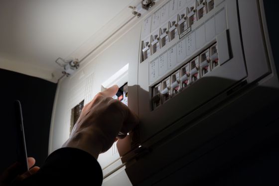 Une main déverrouille un tableau électrique blanc fixé en hauteur sur un mur dans une pièce sombre