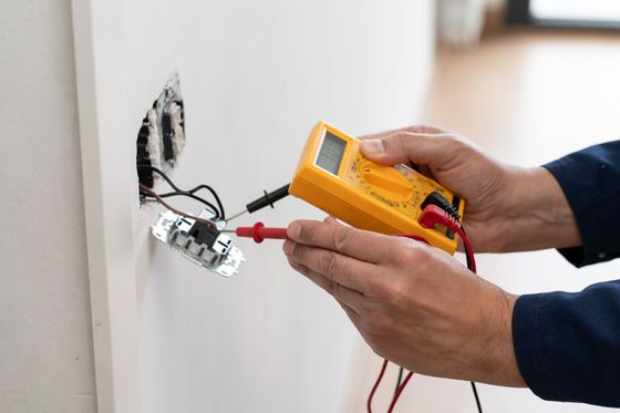 Un technicien vérifie des fils électriques apparents dans le mur à l’aide d’un multimètre, à côté d’un boîtier de prise électrique