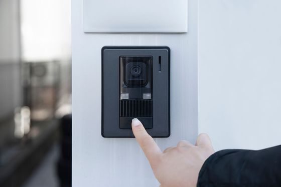 Un doigt appuie sur un bouton d’interphone noir fixé sur un panneau mural blanc