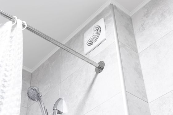 Salle de bains blanche avec douche, barre chromée, rideau, douchette et ventilateur d’extraction sur un mur carrelé