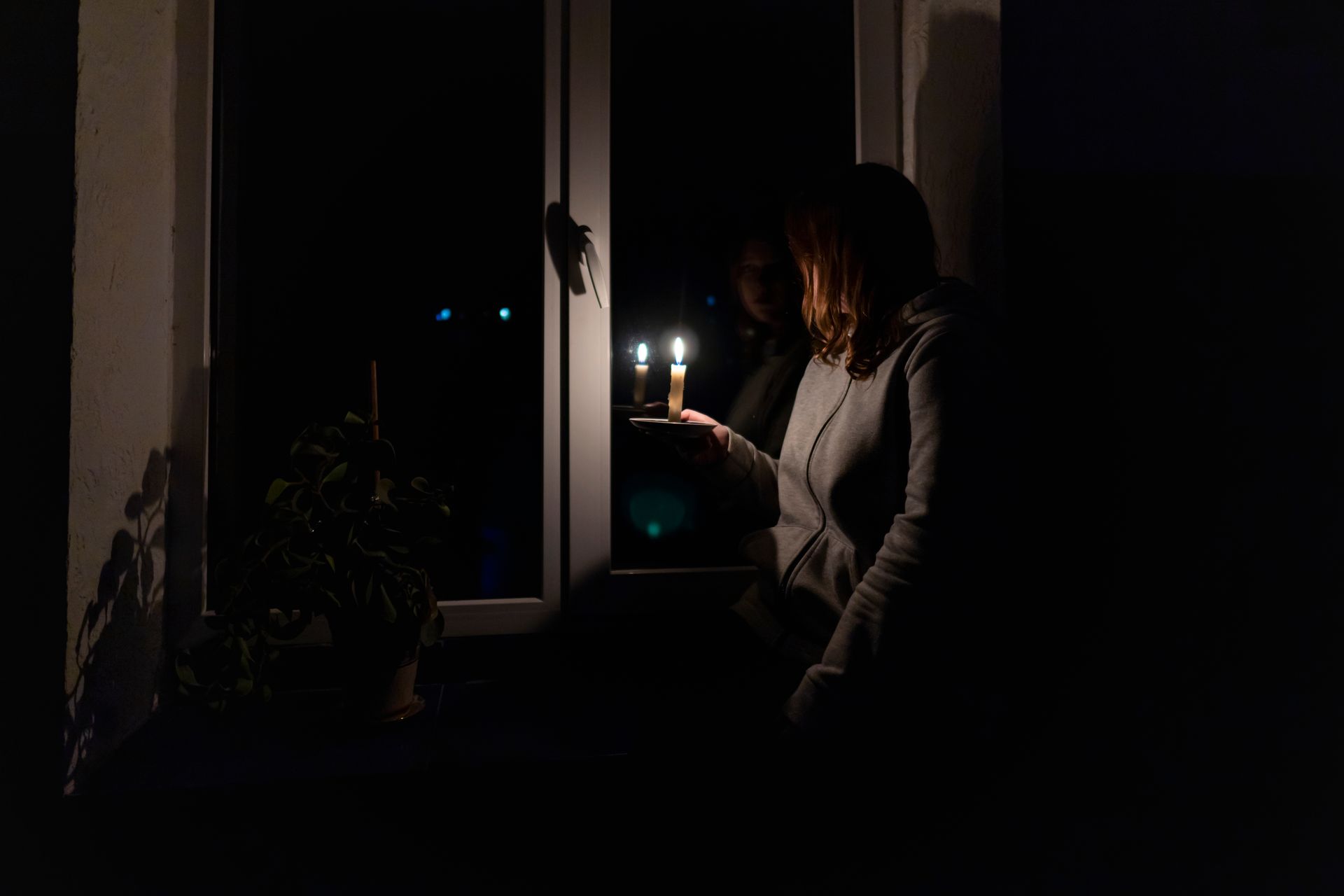Personne tenant une bougie allumée dans une pièce sombre près d’une fenêtre, avec une plante à proximité.