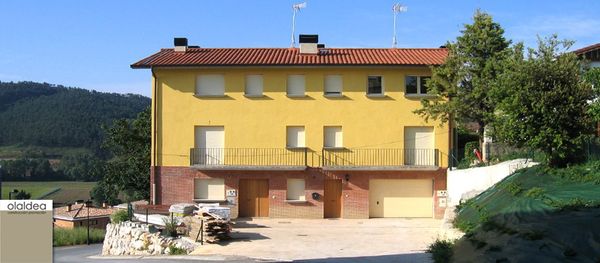 Casa amarilla con techo rojo, garaje y balcón. Con un cielo azul y algunos árboles como fondo.