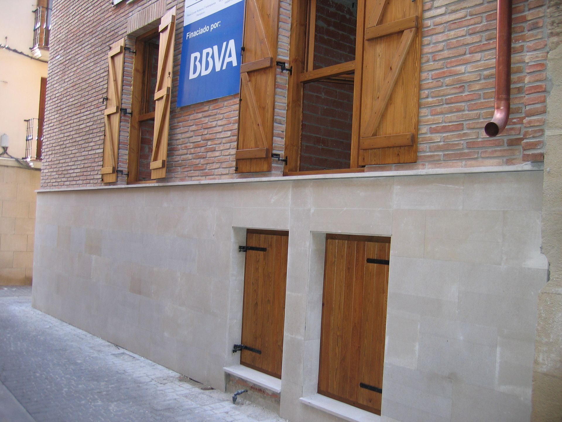 Un edificio con persianas de madera, ventanas y un cartel del BBVA. Persianas marrones, paredes beige.